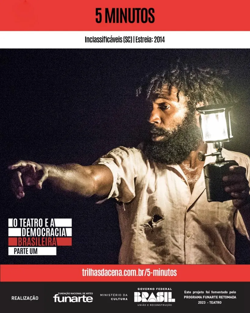 Um homem negro com barba segura uma lanterna, iluminando seu rosto e parte superior do corpo na escuridão. Ele está vestindo uma camisa de cor clara com pequenos rasgos, e aponta para a distância com a outra mão. O texto traz informações sobre o endereço eletrônico da página do espetáculo 5 minutos no Trilhas da Cena. Foto: Rodrigo Sambaqui