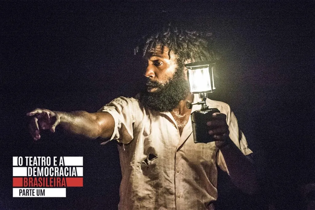 Um homem negro com barba segura uma lanterna, iluminando seu rosto e parte superior do corpo na escuridão. Ele está vestindo uma camisa de cor clara com pequenos rasgos, e aponta para a distância com a outra mão. Foto: Rodrigo Sambaqui