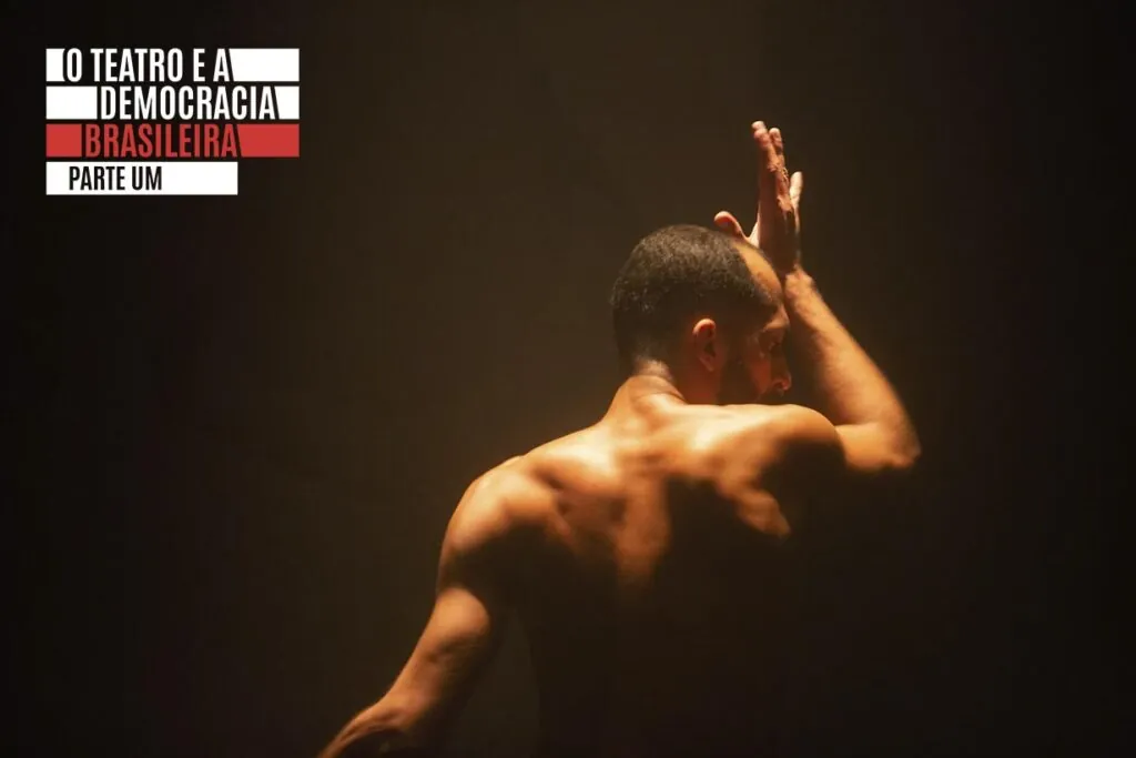 Um homem sem camisa com músculos tonificados está em pé sob iluminação quente e suave em tronco. Ele se encontra de costas, com uma mão tocando a testa. A cena é pouco iluminada, contra um fundo escuro. Foto: Guto Muniz