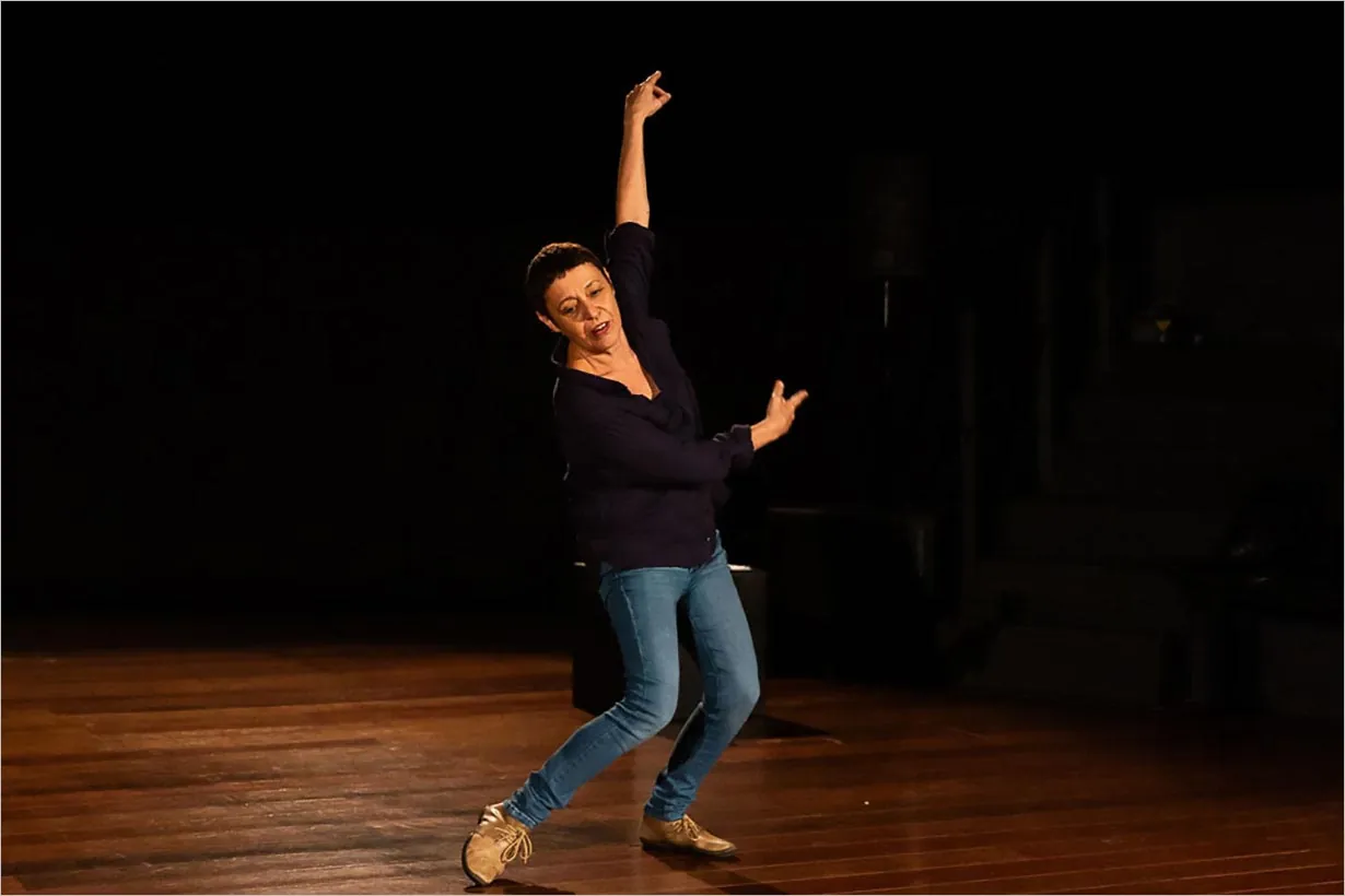 Uma mulher em um palco de madeira faz uma pose de dança com um braço levantado e o outro dobrado, vestindo uma camisa escura, calça jeans azul e sapatos bege, com um fundo escuro atrás dela.