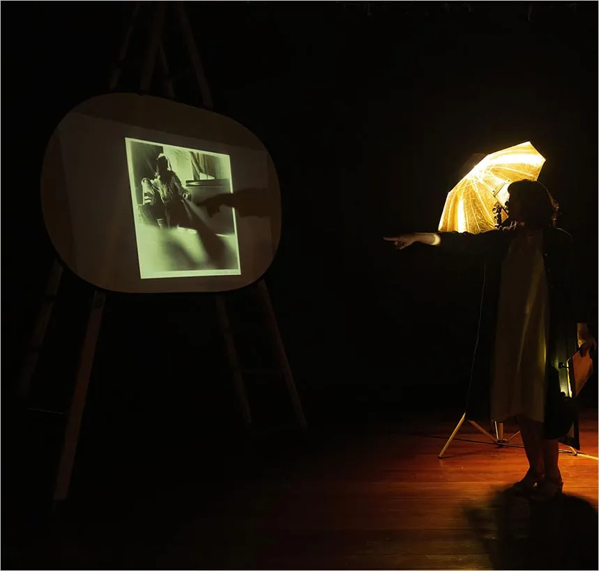Uma pessoa está próxima a uma sombrinha dourada iluminada, apontando para uma foto em preto e branco projetada em uma tela em uma sala pouco iluminada com piso de madeira. Foto: Guto Muniz