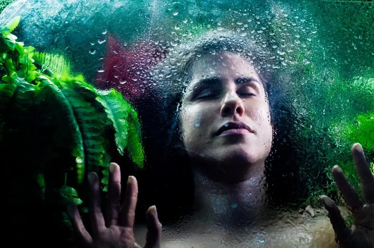 Uma mulher de olhos fechados está deitada sob uma superfície de vidro coberta de gotas d'água. Suas mãos pressionam o vidro, cercada por plantas verdejantes. Foto: Desiree Koppke