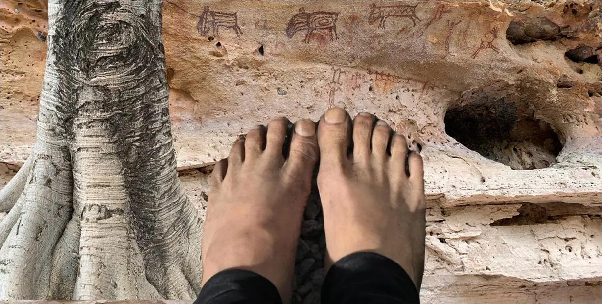 Pés descalços são mostrados em pé sobre um solo rochoso em frente a uma parede de rocha desgastada pelo tempo com esculturas antigas ou pinturas rupestres e um close-up de um tronco de árvore no lado esquerdo da imagem.