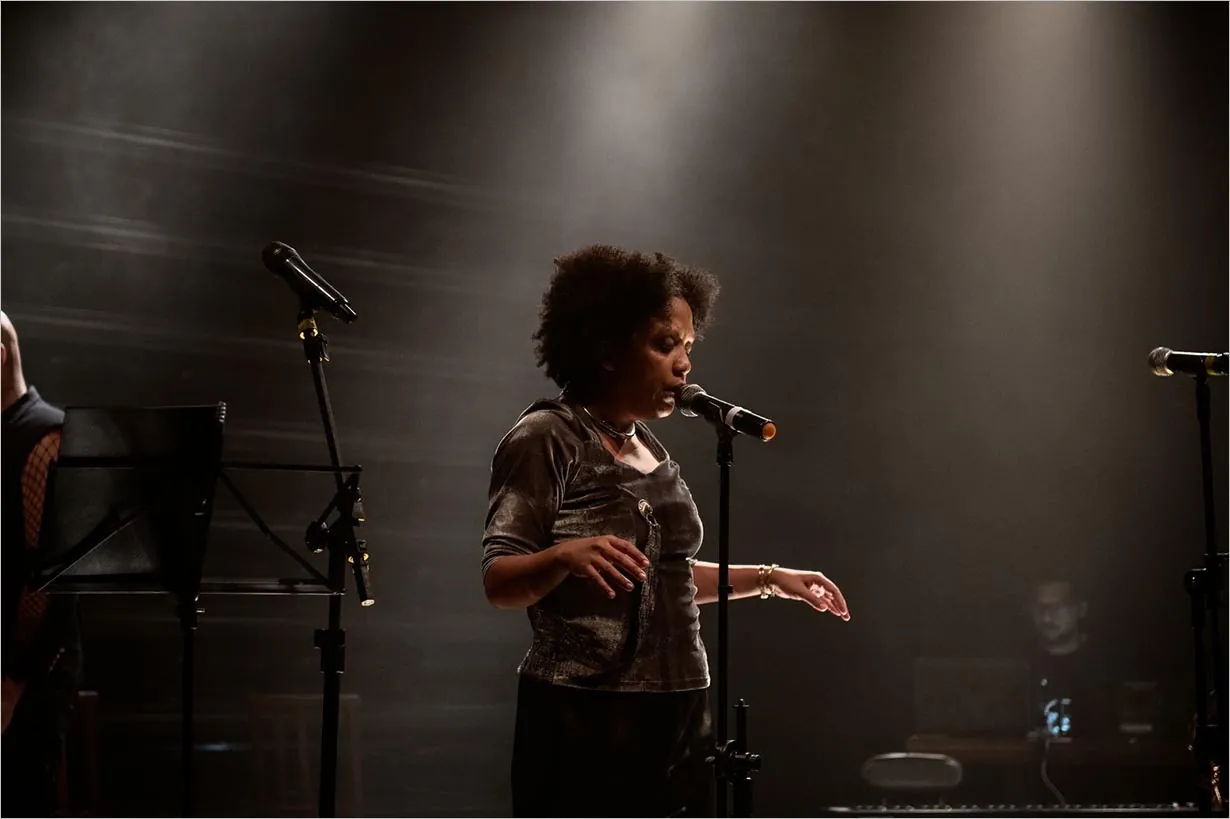Uma mulher canta em um microfone em um palco. Dois outros microfones e um suporte para partituras estão próximos, e uma pessoa trabalha em um laptop ao fundo. Foto Íra Barillo
