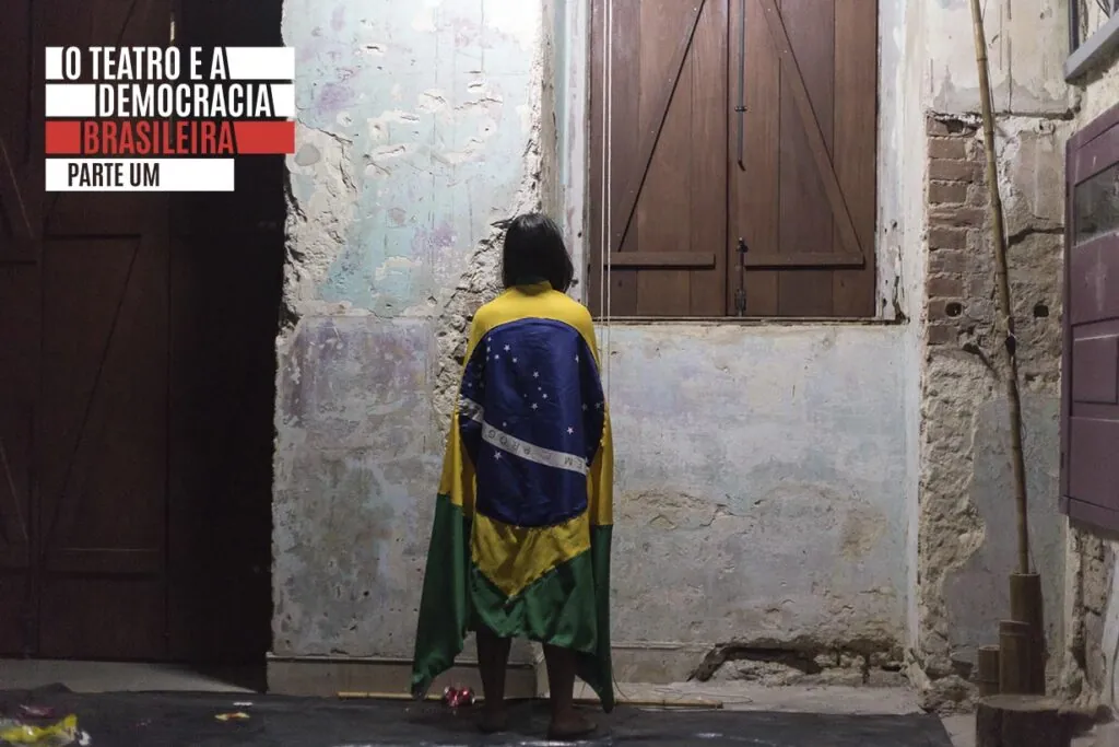 Uma mulher está de costas para a câmera, envolta em uma bandeira brasileira. Ela está de frente para uma parede texturizada com persianas de madeira fechadas, em um espaço com tinta descascada e tijolos aparentes e ásperos. No canto superior esquerdo da imagem está a logo do projeto O teatro e a democracia brasileira. Foto: Adriana Marchiori