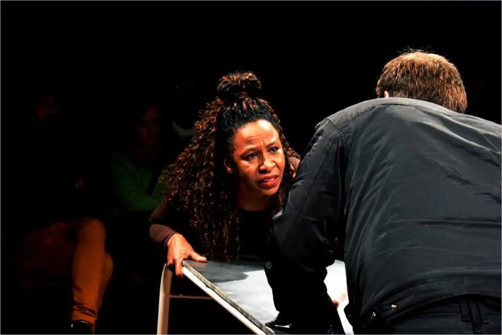 Uma mulher de cabelos cacheados se inclina para frente sobre uma mesa, olhando atentamente para um homem de paletó escuro que a encara de costas para a câmera. O fundo é escuro, com algumas pessoas sentadas pouco visíveis. Foto: Regina Peduzzi Protskof