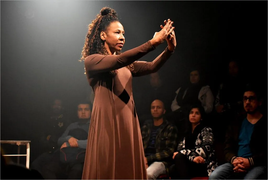 Uma mulher usando um longo vestido marrom está no palco, gesticulando expressivamente com as duas mãos levantadas. Os membros sentados da plateia a observam atentamente no teatro pouco iluminado. Foto: Regina Peduzzi Protskof