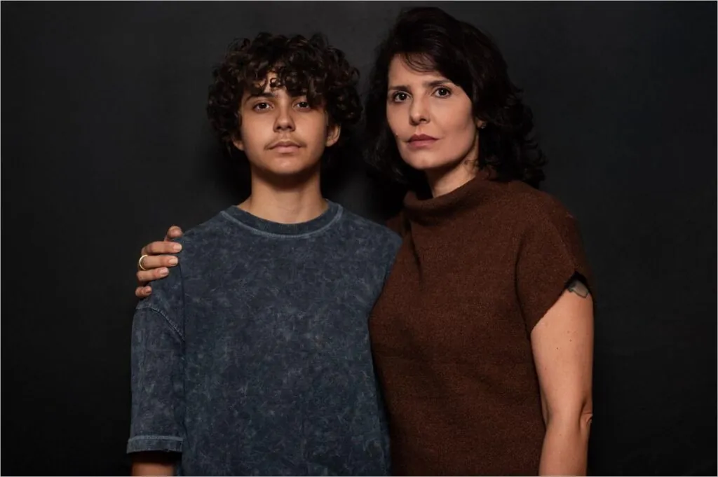 Uma mulher e um adolescente estão juntos contra um fundo escuro. A mulher tem o braço em volta do ombro do menino. Ambos têm expressões sérias e cabelos escuros. Foto: Ana Alexandrino