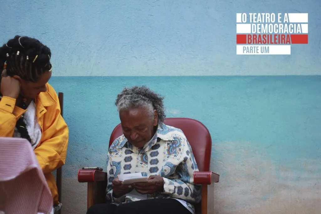 Uma mulher idosa está sentada em uma cadeira vermelha lendo um pedaço de papel, enquanto uma mulher mais jovem ao lado dela, vestida com uma jaqueta amarela, está inclinada com a cabeça apoiada na mão contra uma parede pintada de azul e turquesa. Foto: Tetembua Dandara