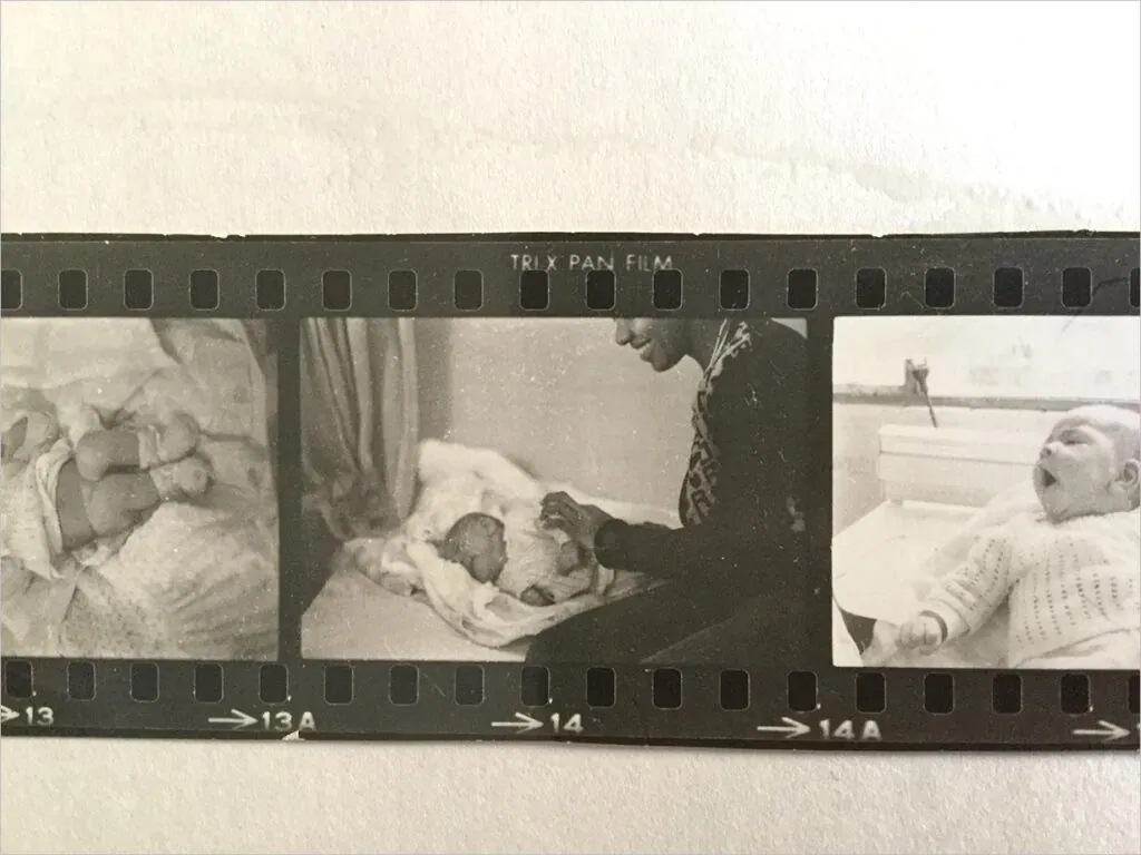 Uma tira de negativos de filme preto e branco captura três fotos: um bebê recém-nascido deitado, uma mulher inclinada sobre o bebê e o bebê bocejando ou chorando em um berço. Foto: Tetembua Dandara