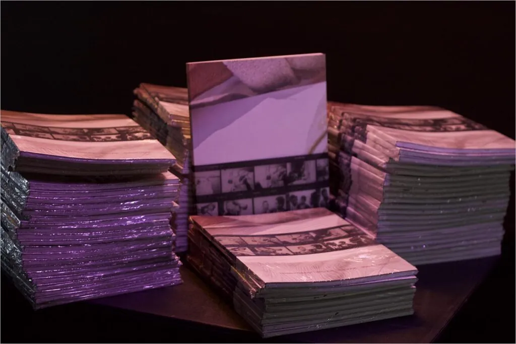 Pilhas de livros com capas brilhantes apresentando imagens de tiras de filme, dispostas sob iluminação roxa sobre um fundo escuro. Foto: Tetembua Dandara