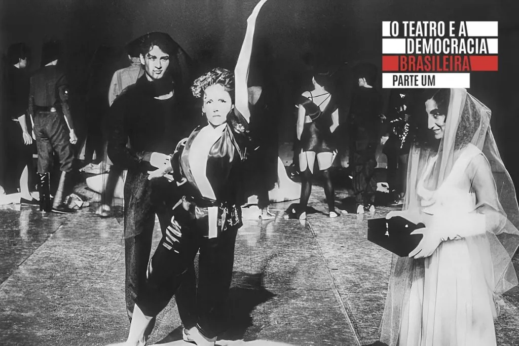 Uma mulher de quimono e touca posa dramaticamente com um braço levantado no palco, ladeada por um homem com roupas escuras e outra mulher com um véu segurando uma caixa, com várias pessoas ao fundo.