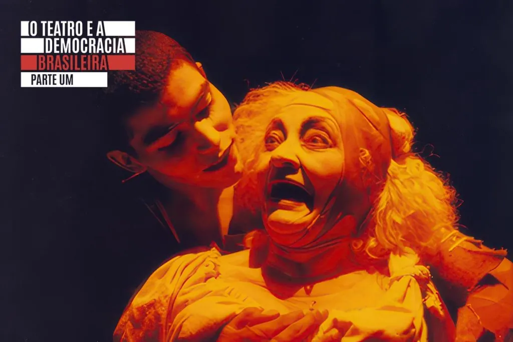 Uma pessoa de cabelos curtos se aproxima de outra pessoa com cabelos loiros encaracolados e maquiagem dramática, que parece estar com os olhos arregalados e gritando. A cena é banhada por uma intensa iluminação laranja, criando um clima dramático e teatral.