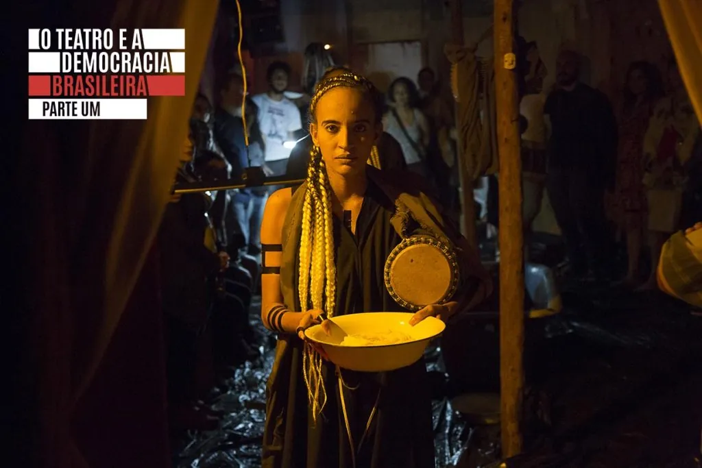Uma mulher em uma sala escura segura uma tigela e um tambor, usando roupas pretas e miçangas. Atrás dela, há pessoas assistindo. O texto no canto superior esquerdo diz: O Teatro e a Democracia Brasileira Parte Um.
