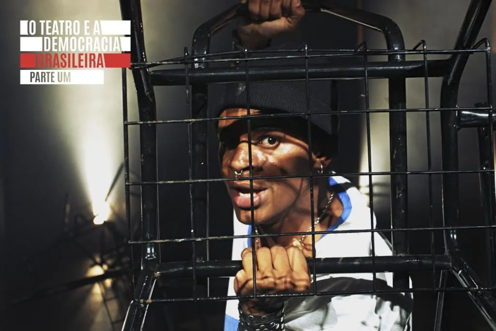 Uma pessoa usando um gorro preto se segura em uma grade de metal que, na verdade, é um compartimento inferior de uma cadeira estudantil, destinado à colocação de materiais escolares. Uma iluminação dramática projeta sombras em seu rosto. Ela está olhando através das grades, com a boca aberta e com uma fisionomia expressiva e enérgica.