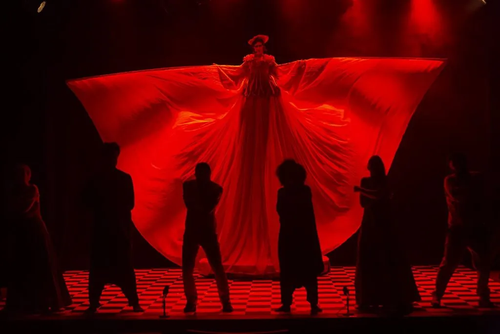 Uma artista em um vestido vermelho largo e dramático está elevada no palco, iluminada por uma luz vermelha, com seis figuras em silhueta abaixo em um piso xadrez.